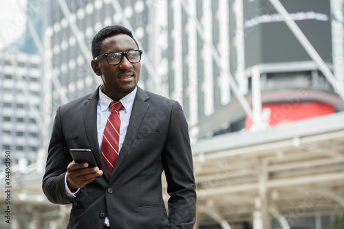 Young happy African businessman holding mobile phone in front of modern building at Bangkok city