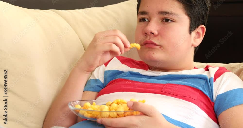 Adolescent boy with acne pimples on the face lying on couch watching tv and eating oily and fryed snacks Adolescent boy with acne pimples on the face lying on couch watching tv and eating oily and fryed snacks