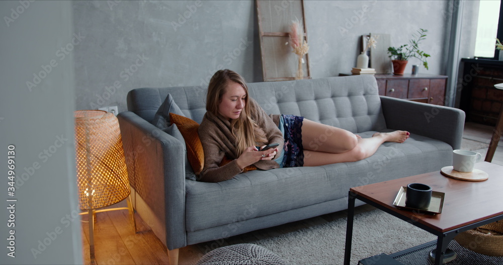 Obraz premium Happy young beautiful Caucasian blonde woman using smart phone at home on cozy sofa, lying down to relax and take a nap.