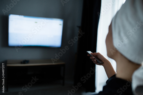 Close-up rear view. A girl in a bathrobe and with a towel on her head, sits on the couch and switches channels on the TV