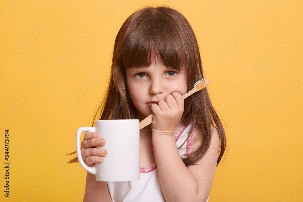 Closeup portrait of cute sad female child wearing t shirt, doing morning dental procedures, looks upset, having toothache, has problems with milk tooth, posing isolated over yellow studio background. Closeup portrait of cute sad female child wearing t shirt, doing morning dental procedures, looks upset, having toothache, has problems with milk tooth, posing isolated over yellow studio background.