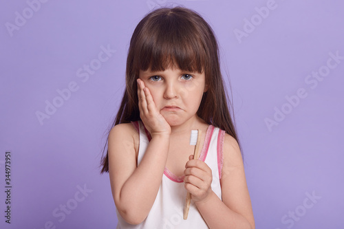 Studio shot of upset little girl posing isolated over lilac studio background, keeping palm on cheek, looks sad, has toothache, child's first teeth fall, kid holds tooth brush in hand, looks at camera