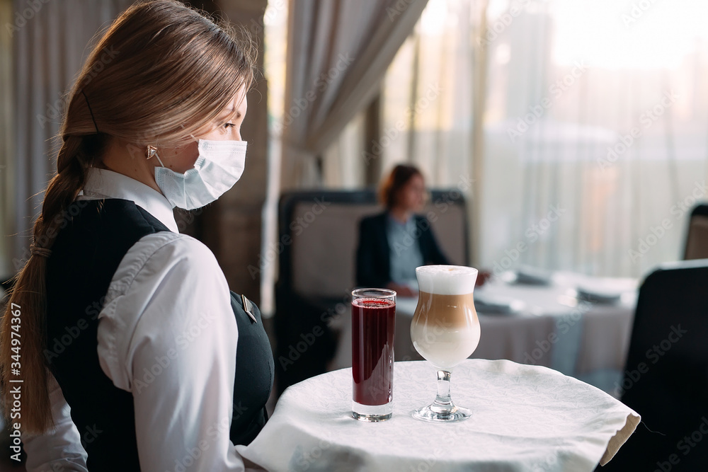 A female Waiter of European appearance in a medical mask serves Latte coffee. A female Waiter of European appearance in a medical mask serves Latte coffee.