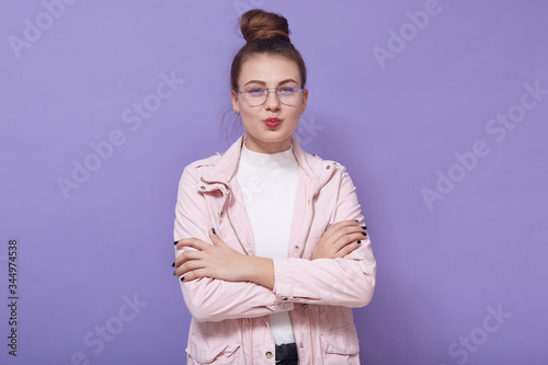 Portrait of beautiful magnetic lovely teen looking directly at camera, having pleasant facial expression, having folded arms, crossing hands, standing isolated over lilac background in studio.