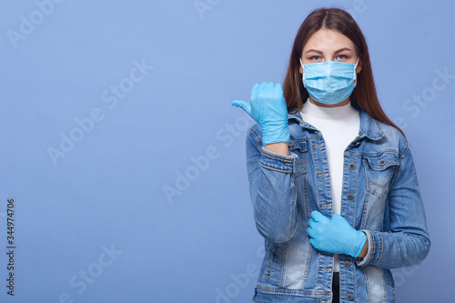 Indoor photo of confident attractive young woman looking directly at camera, making gesture, showing direction with thumb, wearing gloves and antibacterial mask. Copyspace for advertisement. photo