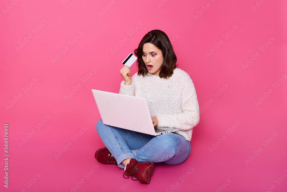 Indoor shot of woman sitting on floor, wearing casual attire, having astonished facial expression, keeps mouth opened, holding credit card in hand, doing online shopping, being shocked from prices. Indoor shot of woman sitting on floor, wearing casual attire, having astonished facial expression, keeps mouth opened, holding credit card in hand, doing online shopping, being shocked from prices.