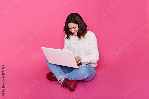 Quadro su tela Horizontal shot of smiling brunette lady sitting on floor with crossed legs and holding white lap top,looking at its screen with smile, posing isolated over pink background, using wireless Internet