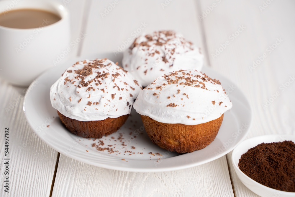 Cupcakes with whipped cream and chocolate chips on a white plate and a Cup of coffee in the background, on a white wooden table. Image for the menu or catalog of confectionery products. Cupcakes with whipped cream and chocolate chips on a white plate and a Cup of coffee in the background, on a white wooden table. Image for the menu or catalog of confectionery products.