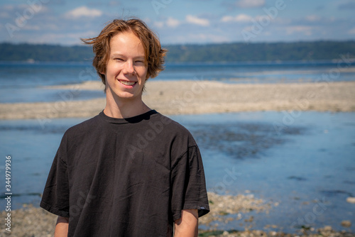 A smiling handsome teenage boy stands on a beach in the Pacific Northwest and poses for a portrait picture.