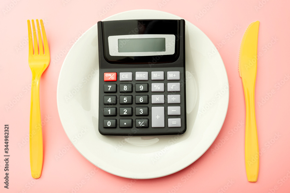 Calculate expensive food spending costs, counting calories and weight loss program concept with calculator onn empty plate, yellow fork and knife isolated on pink background