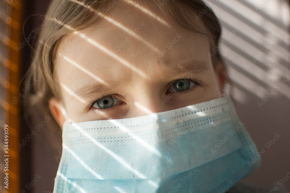 Coronavirus Covid-19 outbreak. Little blonde girl with blue eyes wearing disposable mask for protection of virus sitting near the window with blinds. Personal protective equipment Coronavirus Covid-19 outbreak. Little blonde girl with blue eyes wearing disposable mask for protection of virus sitting near the window with blinds. Personal protective equipment