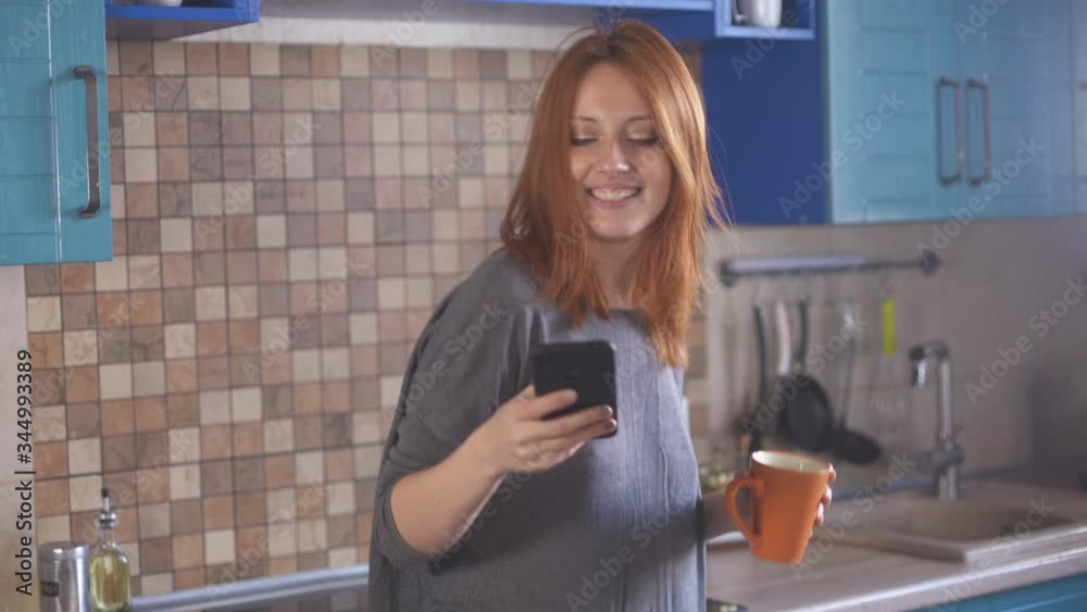 Girl with red hair in morning in kitchen listens to music, sings and dances. Young redhead woman dancing with a cup of coffee and a phone in her hands. Happy hipster millennial girl in t-shirt. Girl with red hair in morning in kitchen listens to music, sings and dances. Young redhead woman dancing with a cup of coffee and a phone in her hands. Happy hipster millennial girl in t-shirt.