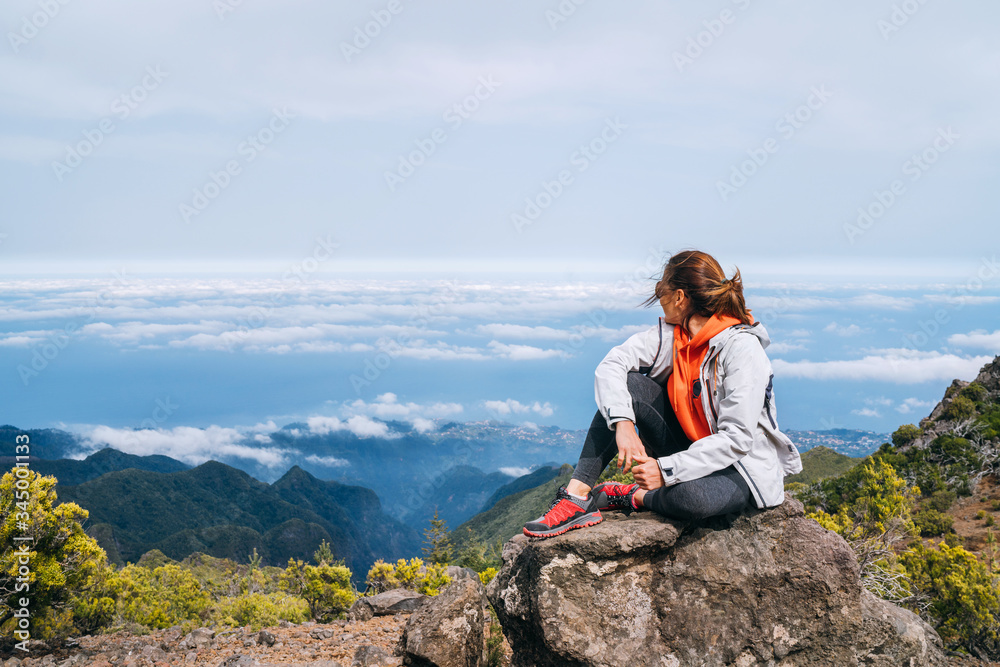 Young adult caucasian female dressed  sporty clothes resting on the rock and enjoying clouds view from Pico Ruivo mount 1861m - the highest peak on the Madeira Island,Portugal. Active vacation concept