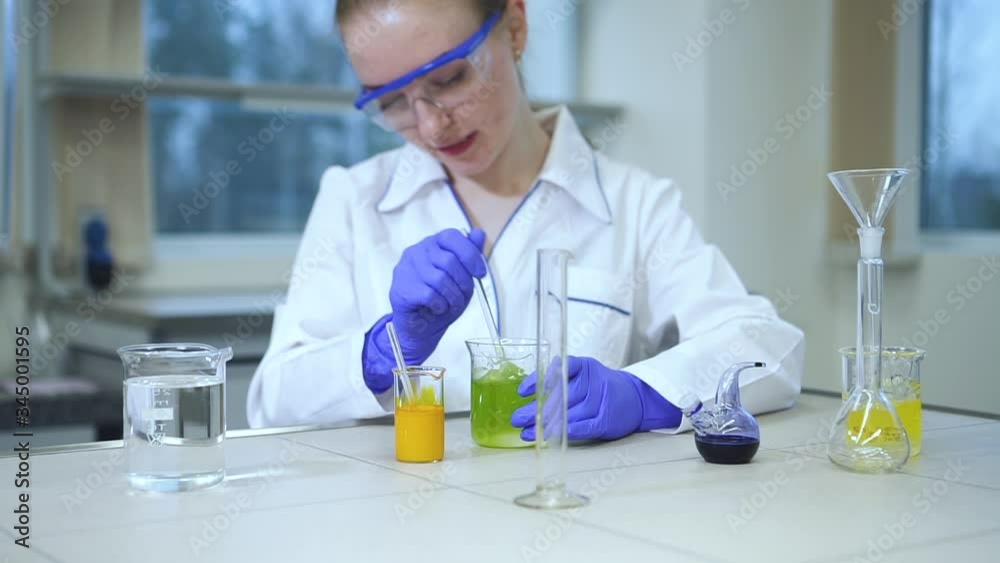 Lab technician is in work process with chemicals, sitting at desk in pharmaceutical laboratory spbd. Woman employee stirs colored liquid in flask with glass stick at workplace in company. Modern Lab technician is in work process with chemicals, sitting at desk in pharmaceutical laboratory spbd. Woman employee stirs colored liquid in flask with glass stick at workplace in company. Modern