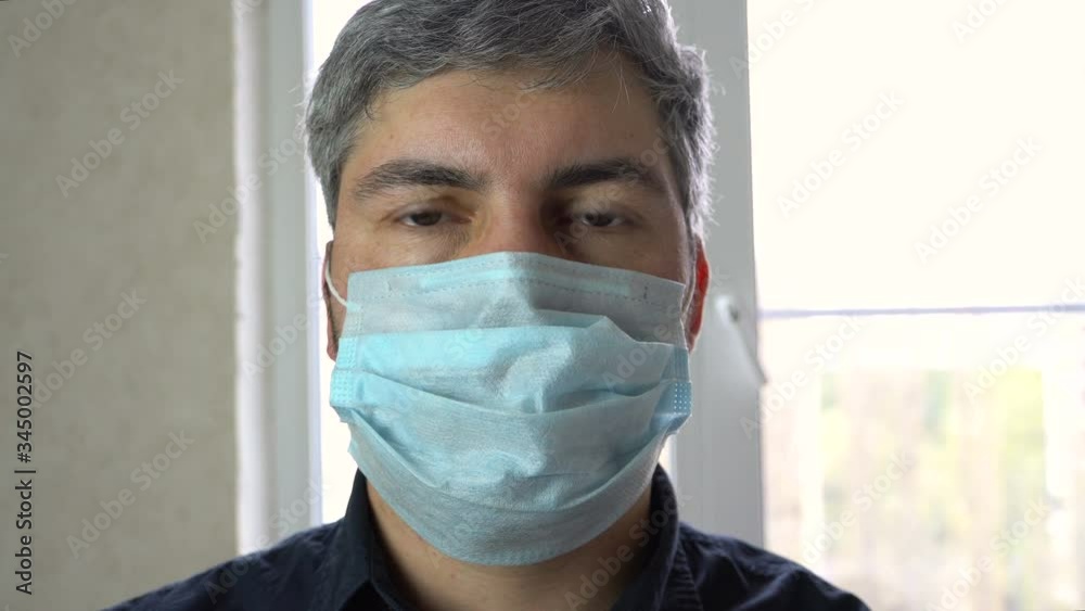 A man in a medical mask stands at the window. Dark shirt. Sick person. Protective equipment. Quarantine. Close-up. A man in a medical mask stands at the window. Dark shirt. Sick person. Protective equipment. Quarantine. Close-up.