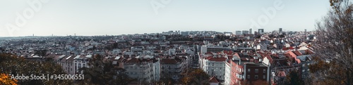 A panoramic shot of a Lisbon from high above: an urban skyline with plenty of traditional Portuguese houses, streets, and roads; a row of trees in a defocused foreground, sunny day