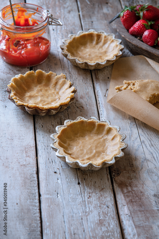 Cooking process and ingredients for the recipe of tartlets with berry cream. Raw shortbread base in ceramic baking pans on wooden table. Recipe or cookbook concept. Vertical format, copy space