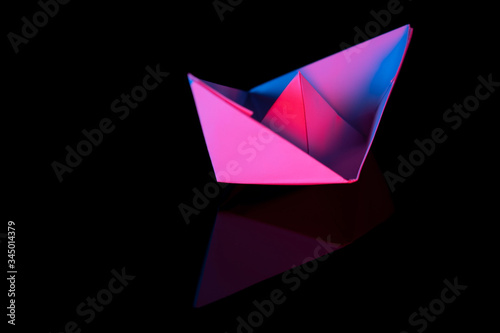 Folded white paper boat, isolated on black background. Illuminated blue and red, with reflection. Close-up with selective focus.