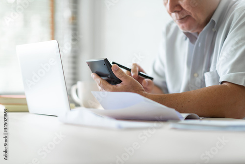 Stressed Asian Old man using calculator for calculate family budget, monthly expenses in home during economic crisis. Senior male worry about his Debts with account book, bill, passbook, receipt