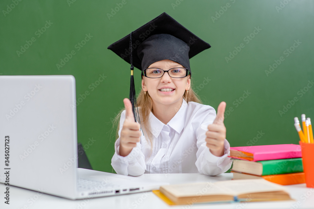 Happy smart girl wearing a graduation cap uses laptop near the green school chalkboard and shows thumbs up gesture Happy smart girl wearing a graduation cap uses laptop near the green school chalkboard and shows thumbs up gesture