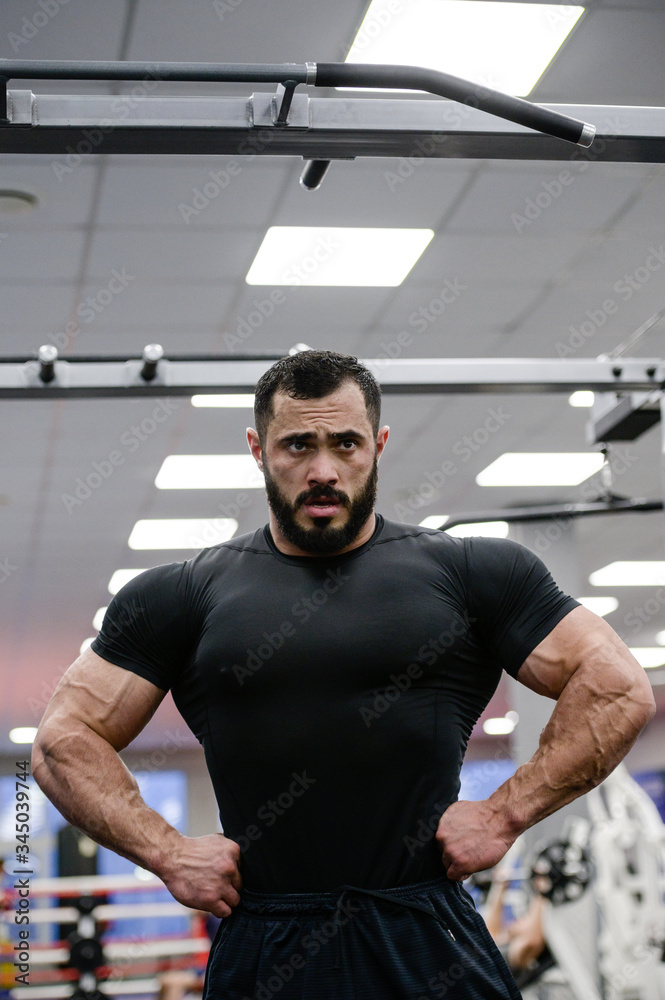 strong young man with beard wearing black jersey showing his big muscles in sport gym during workout posing strong young man with beard wearing black jersey showing his big muscles in sport gym during workout posing