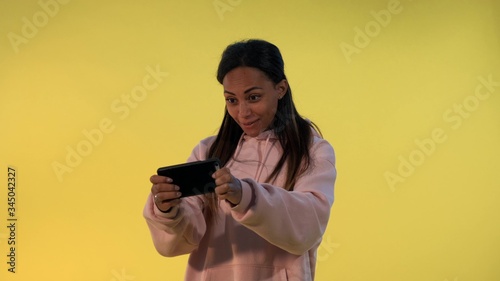 Pleasant-looking black girl playing games on smartphone on yellow background. She is passionate about the game.