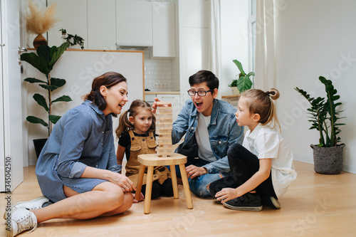 Dad nerviously placing wooden brick on top while kids, mom wait for him to fail