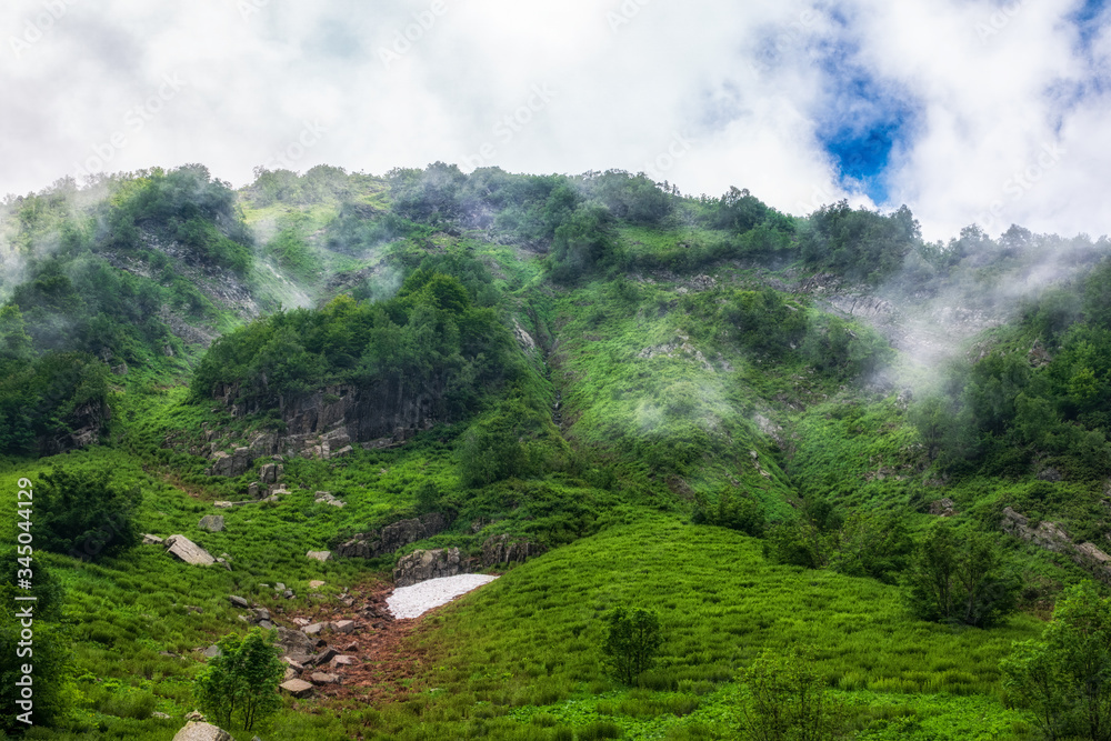 High mountain with green slopes, rocks and not melted snow hidden in thick clouds and fog.