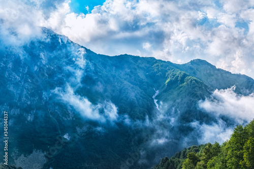 High mountains with green slopes, rocks and not melted snow hidden in thick clouds and fog.