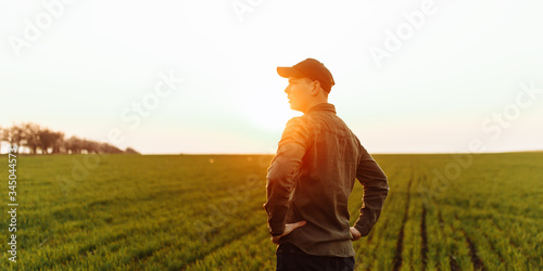Young farmer stands in the green field checking and waiting for harvest to grow. Sunset view of a boy wearing green shirt and a cap walking on the lawn. Agricultural concept. © Konstantin Zibert