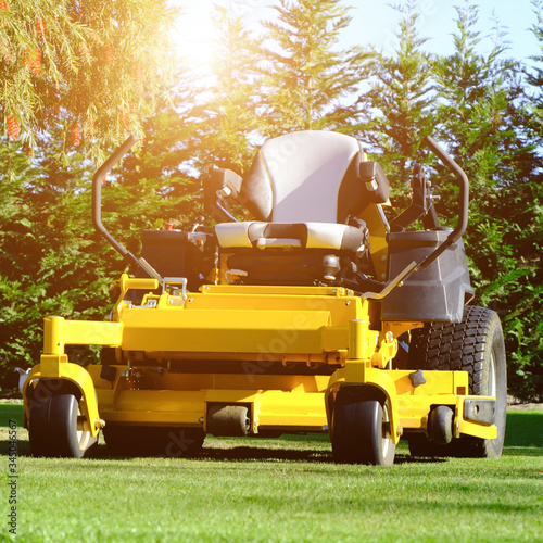 The yellow zero-turn mower parked in the middle of the green grass field, the orange sunlight splashes behind and the green background fence, mow the lawn service concept.