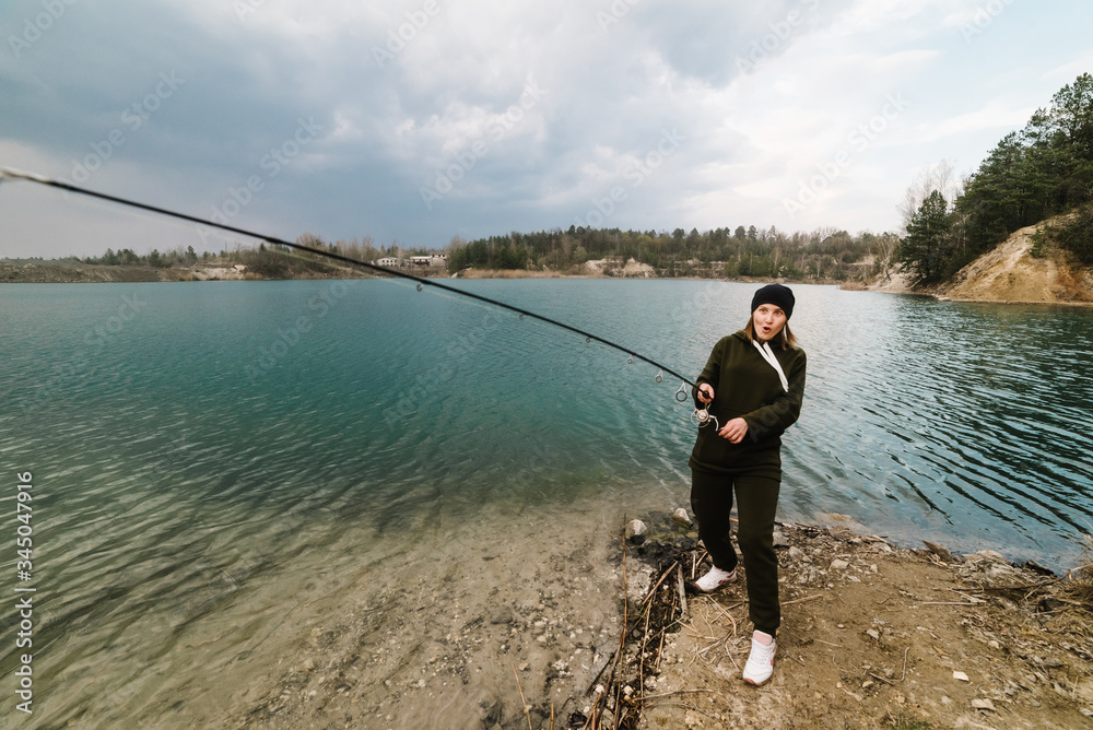 Fisherman with rod, spinning reel on the river bank. Fishing for pike, perch, carp. Woman catching fish, pulling rod while fishing at the weekend. Girl fishing from beach lake or pond with text space.