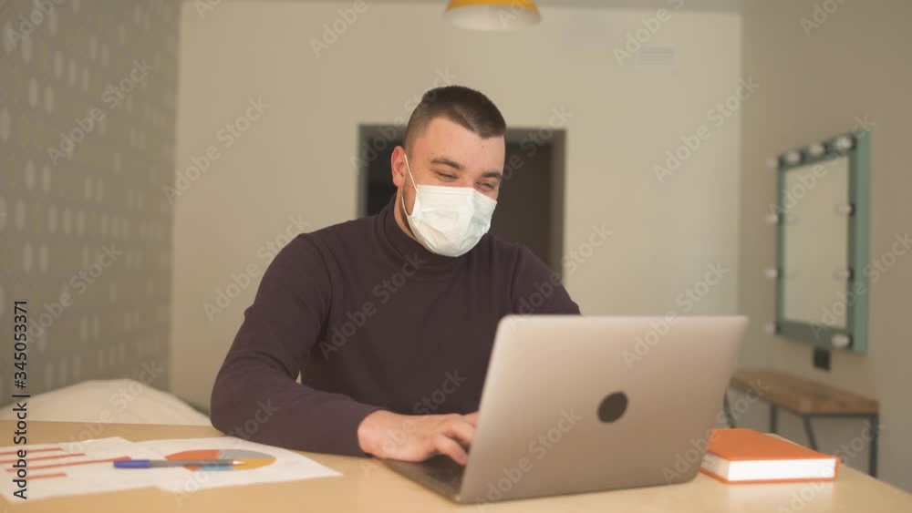 Young businessman in medical mask and brown sweater becomes very happy after seeing his results on a laptop during the pandemic covid-19 coronavirus quarantine. People have to work and study from home