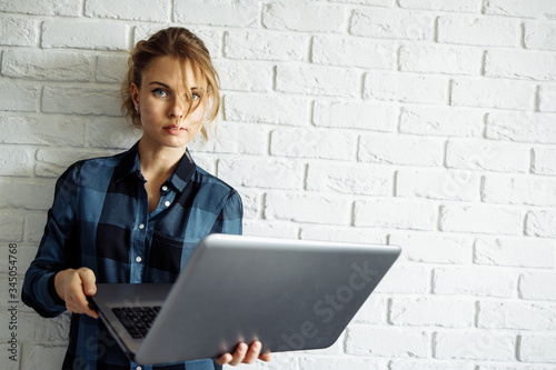 Young woman freelancer with laptop in her hands standing against white brick wall.