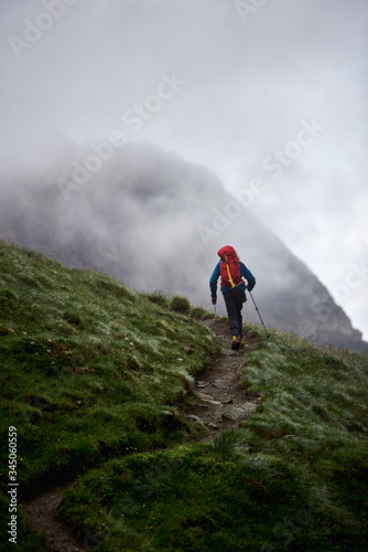Back view of man backpacker with trekking sticks climbing the grassy hill. Traveler with backpack walking on path and heading to foggy mountain. Concept of hiking, climbing and backpacking.