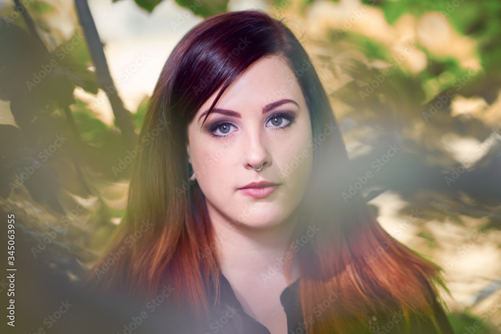 Young woman with gray eyes and an ombre hairstyle in color tones of crimson red and orange, blending in between green dreamy forest foliage with a creamy bokeh. Young woman with gray eyes and an ombre hairstyle in color tones of crimson red and orange, blending in between green dreamy forest foliage with a creamy bokeh.