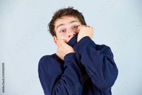 Scared handsome young man pulling up his blue sweater covering his mouth with a dramatic facial expression in the times of the corona pandemic.