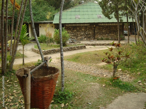 Rattan wastebasket in the yard among palm trees. In the background is a hut with a green roof and tropical plants. Clean, well-kept yard. The flowerbed is lined with smooth stones. Neat paths.