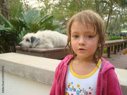 A girl in a pink sweater is talking. In the background on the bench is a large white dog with dark ears. A child and a huge dog.