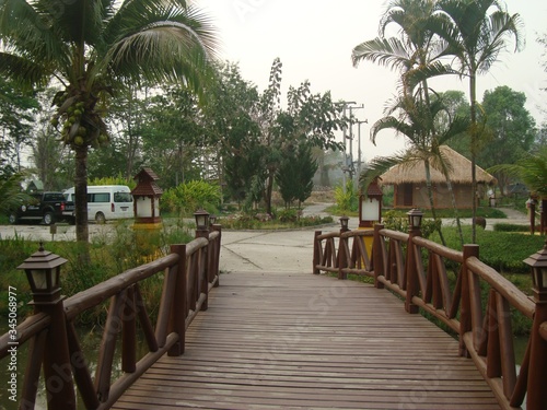 Beautiful wooden bridge over the river. Lanterns on the railing. In the background are palm trees and a hut and cars. Tropical greenery. Travel.