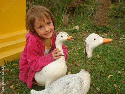A child and a pair of white geese. Old sculptures of geese. A girl in a pink blouse hugs a bird. In the background is a yellow building and green grass.