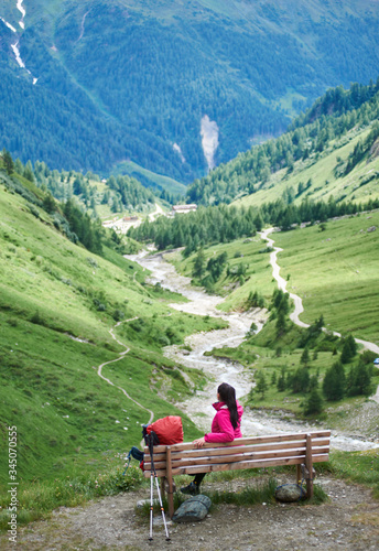 Back view of young woman tourist sitting on bench. Female traveler resting at hillside meadow and enjoying the view of majestic mountains. Concept of travelling, tourism and hiking.