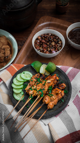 chicken satay on striped napkin and wooden tray also a bowl of sauce and slice of cucumber