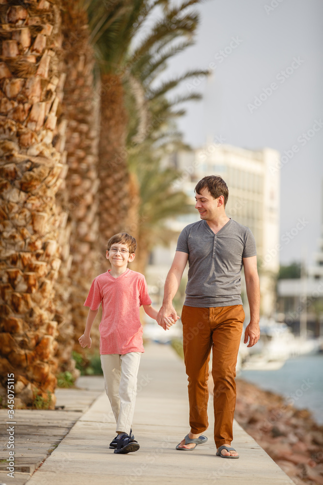 Son & father walking on a beach near palm trees weared in casual clothes. Tropic vacation. Sea relax and rest. Spending a time on a beach. Smiling boy with his Dad together. Summer holiday sunny day Son & father walking on a beach near palm trees weared in casual clothes. Tropic vacation. Sea relax and rest. Spending a time on a beach. Smiling boy with his Dad together. Summer holiday sunny day