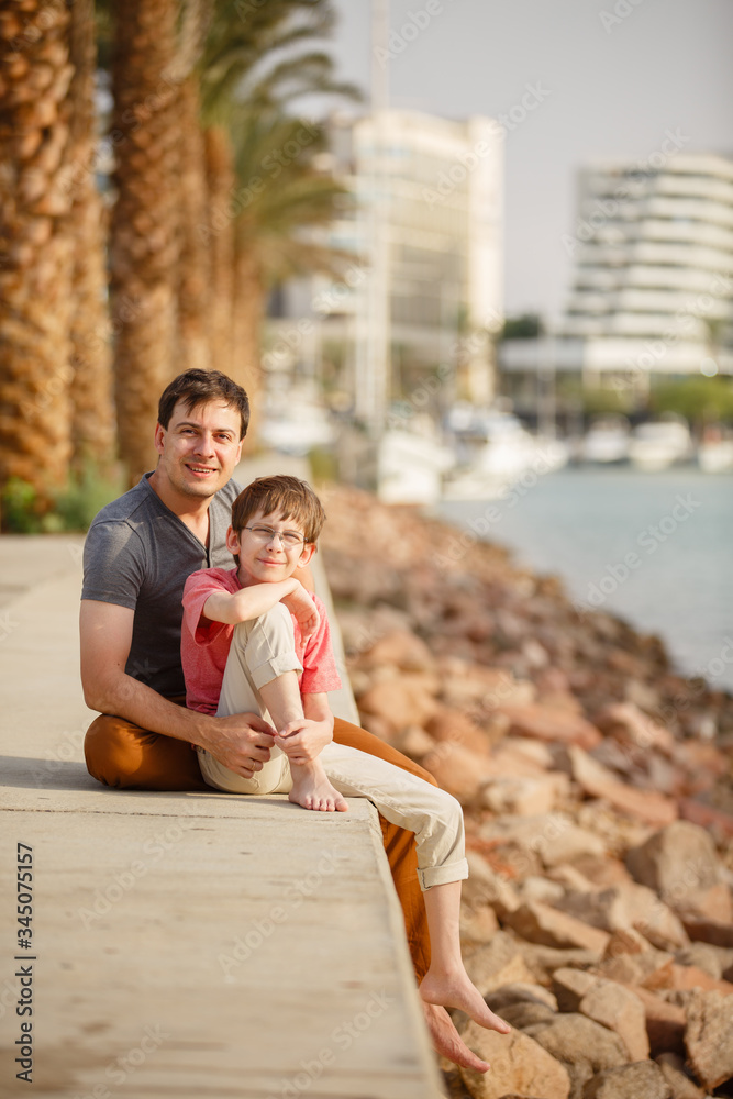 Son & father walking on a beach near palm trees weared in casual clothes. Tropic vacation. Sea relax and rest. Spending a time on a beach. Smiling boy with his Dad together. Summer holiday sunny day