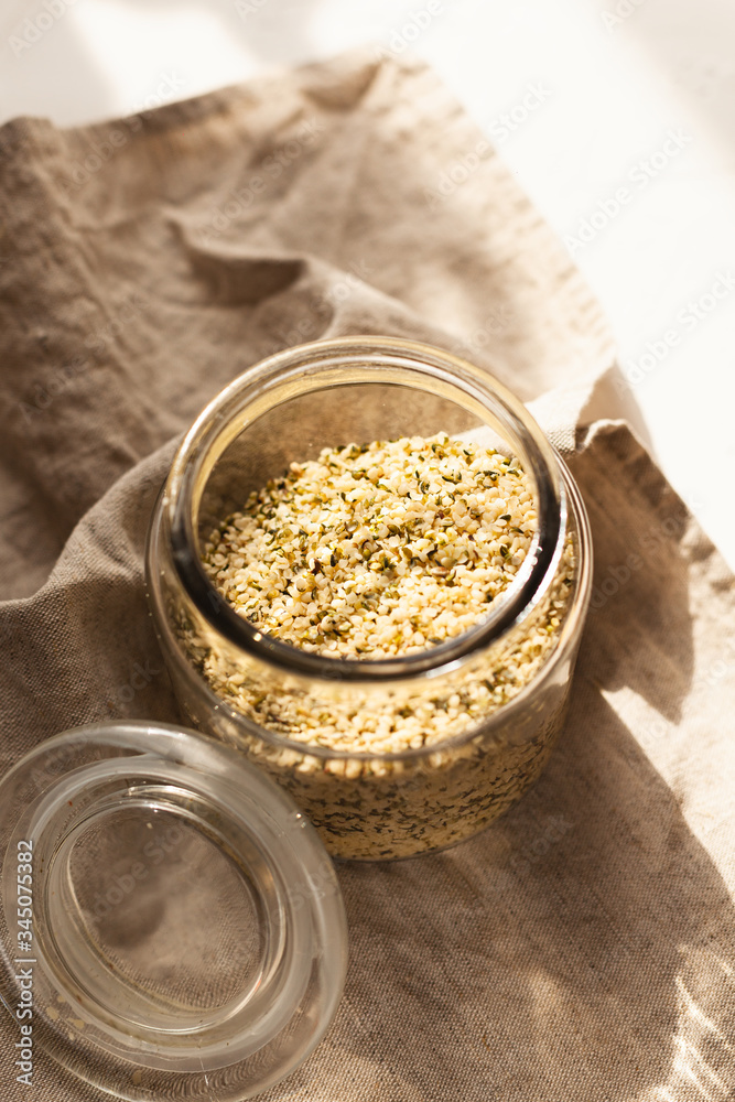Cannabis kernels in a glass jar on a white table with a wooden spoon Cannabis kernels in a glass jar on a white table with a wooden spoon
