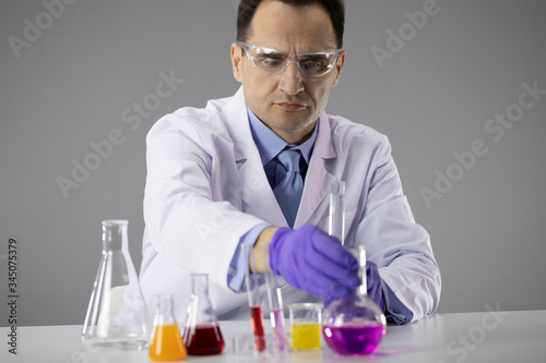 40s Male doctor or Chemist researcher in blue gloves and protective glasses conducting experiments with flasks filled with colorful chemical solutions isolated on grey background. Laboratory concept
