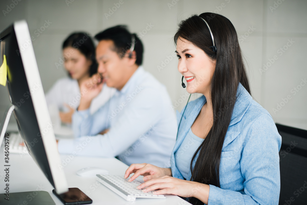 Young beautiful Asian operator woman with headsets working with team in call center.Customer support phone looking computer with smiling and feeling happy.Team operator call center service concept. Young beautiful Asian operator woman with headsets working with team in call center.Customer support phone looking computer with smiling and feeling happy.Team operator call center service concept.