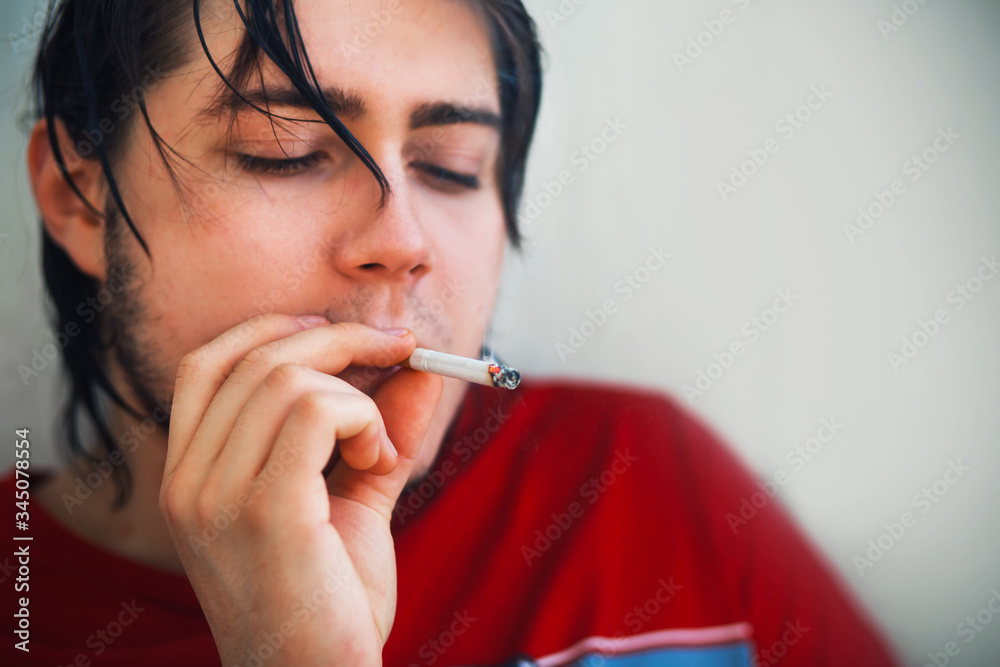 A young handsome guy in a red t-shirt with wet hair after a shower is enjoying smoking a cigarette in the early morning. A young handsome guy in a red t-shirt with wet hair after a shower is enjoying smoking a cigarette in the early morning.