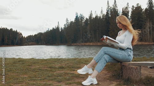 Beautiful young blonde woman sitting and reading book in a park near the lake. Side view